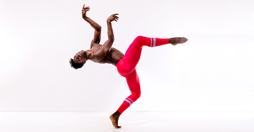 Lloyd Knight is a tall, muscular Black man here wearing red leggings with silver bands at the calves. He poses against a white backdrop in forced arch, the leg closest to the camera raised in parallel attitude front. His torso arches back, nearly parallel to the ground, as he raises his arms in a purposefully bent third position. His head turns toward the camera with an easy, open smile as he tips off-balance.