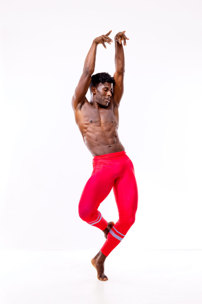 Lloyd Knight, a tall, muscular Black man, balances on forced arch in coupé back. His arms are extended overhead as he hinges at the hips. He wears bright red leggings with silver bands at the calves.