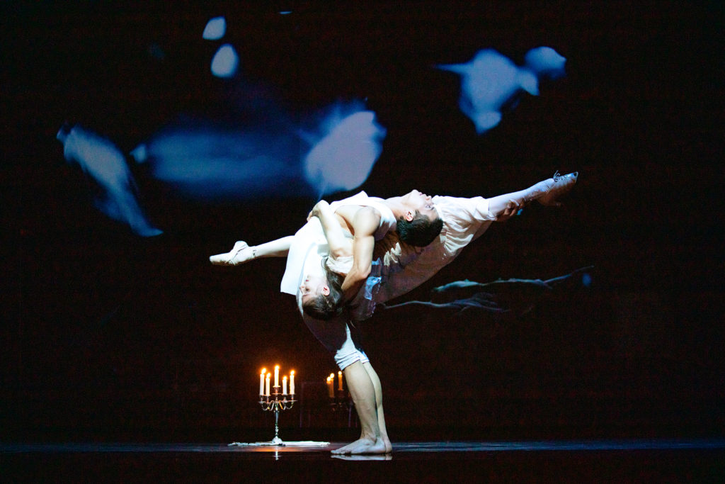 A male dancer stands in profile to the viewer, arching back so his head is parallel with the floor. A ballerina wraps around his back. She is upside down, her legs stretching into a split that is parallel to the floor. Her arms wrap around her partner's torso, while he braces one arm behind her back, the other beneath her back leg. A small lit candelabra sits upstage.