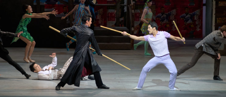 Violence erupts onstage, two male dancers at the center squaring off. One wears a dramatic black duster and wields a long staff; the other wears lilac pants and a matching shirt and holds a shorter wooden stick in each hand, one of which he brandishes towards his enemy's face. The outfits and decor are evocative of 1960s Hong Kong.