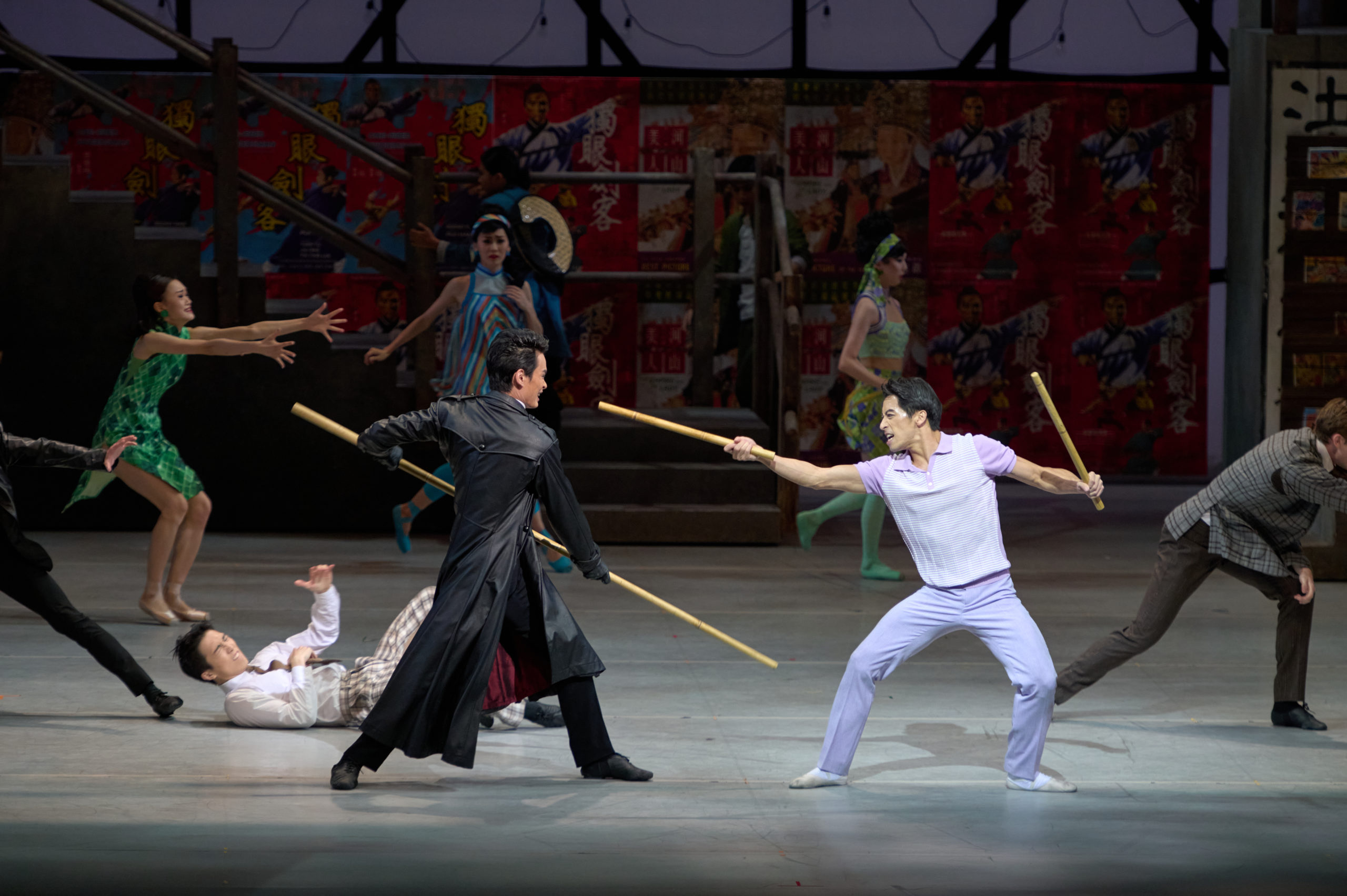 Violence erupts onstage, two male dancers at the center squaring off. One wears a dramatic black duster and wields a long staff; the other wears lilac pants and a matching shirt and holds a shorter wooden stick in each hand, one of which he brandishes towards his enemy's face. The outfits and decor are evocative of 1960s Hong Kong.
