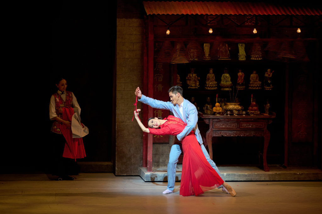 A male dancer in a blue suit dips his partner, a woman in a long red dress and pointe shoes; the fashion is decidedly mid-century. They each hold the ends of a short red ribbon. Upstage is a small shrine.