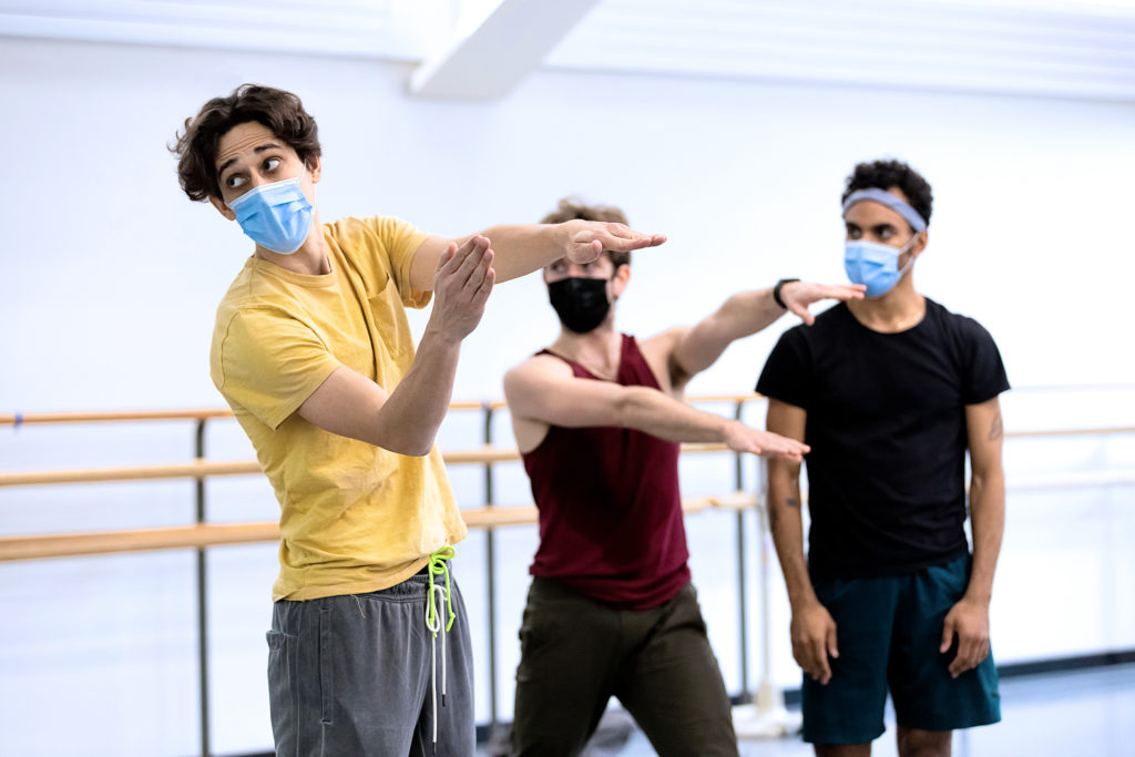 Justin Peck glances over his shoulder toward the front of the room as he demonstrates a gesture to two dancers in the studio just behind him. All wear rehearsal gear and masks over their noses and mouths.