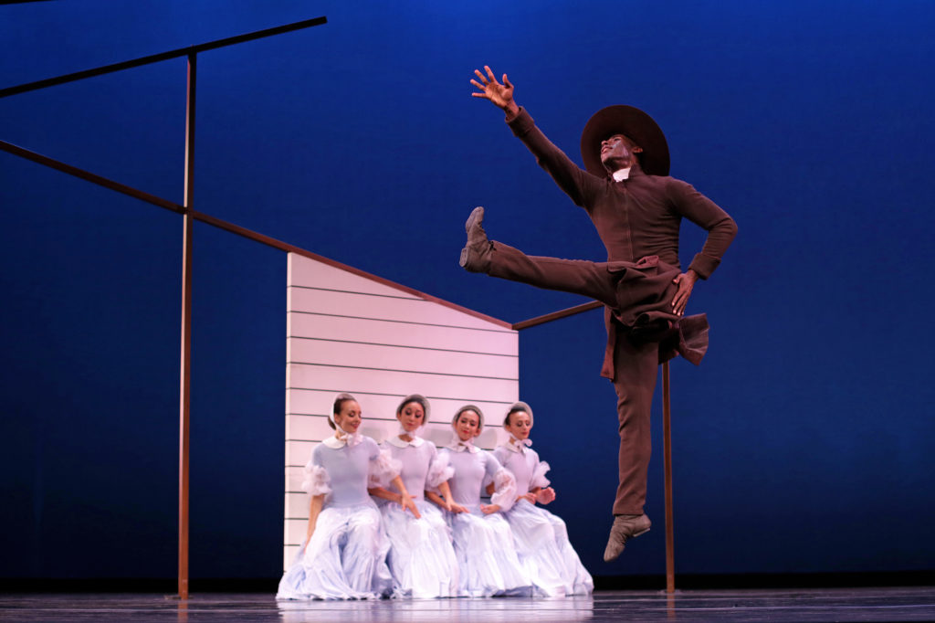 Lloyd Knight is caught mid-jump, a flexed foot extended in front of him at 90 degrees while his opposite arm reaches up and to the corner. He is costumed all in black, with a white preacher's collar and a broad-brimmed black hat. Behind him, four women in period appropriate white bonnets and dresses sit against the set suggesting a house.