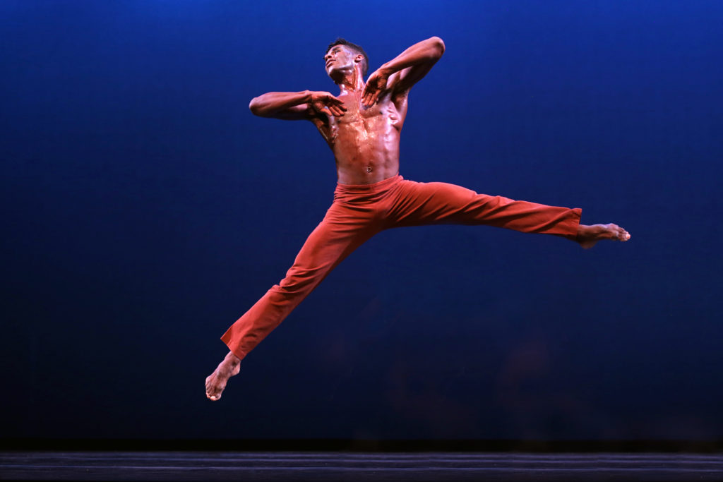 Lloyd Knight is shown mid-jump, legs splitting unevenly to the side. The backs of his hands touch his chest, elbows raised. He looks up and over his right shoulder. He wears orange-red pants.