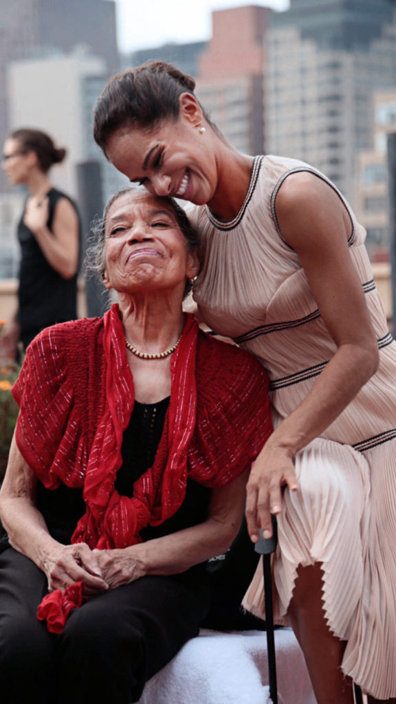 Misty Copeland perches beside a seated Raven Wilkinson, both smiling. Copeland tips her cheek against Wilkinson's forehead, her arm resting against the older woman's back.