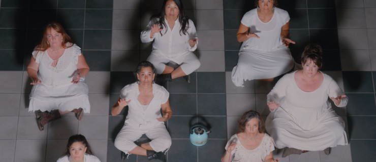 Seven women of various ages, ethnicities and body types sit on the floor and look up at the camera. Their hands are raised as they perform body percussion. All wear white. At the center is a teal blue kettle that has clearly seen use. Their shadows reaching back along the floor, a chessboard pattern of grey and white tiles.