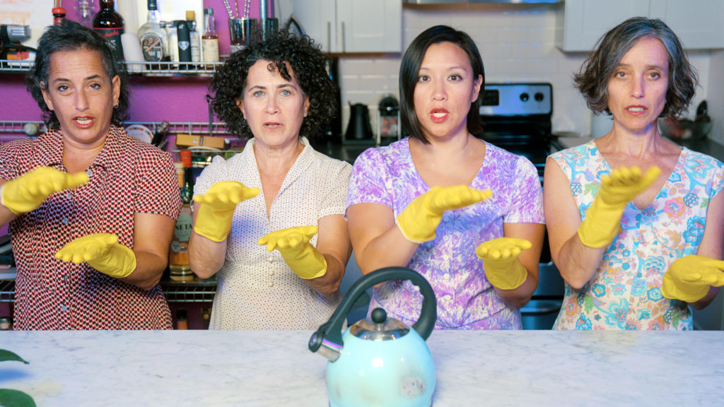 Four women stand behind a kitchen counter where a worn blue kettle rests. Their hands are covered by yellow rubber gloves and are blurred as they move through the shape of a box. The women wear patterned blouses; they gaze at the camera, mouths open mid-speech or song.
