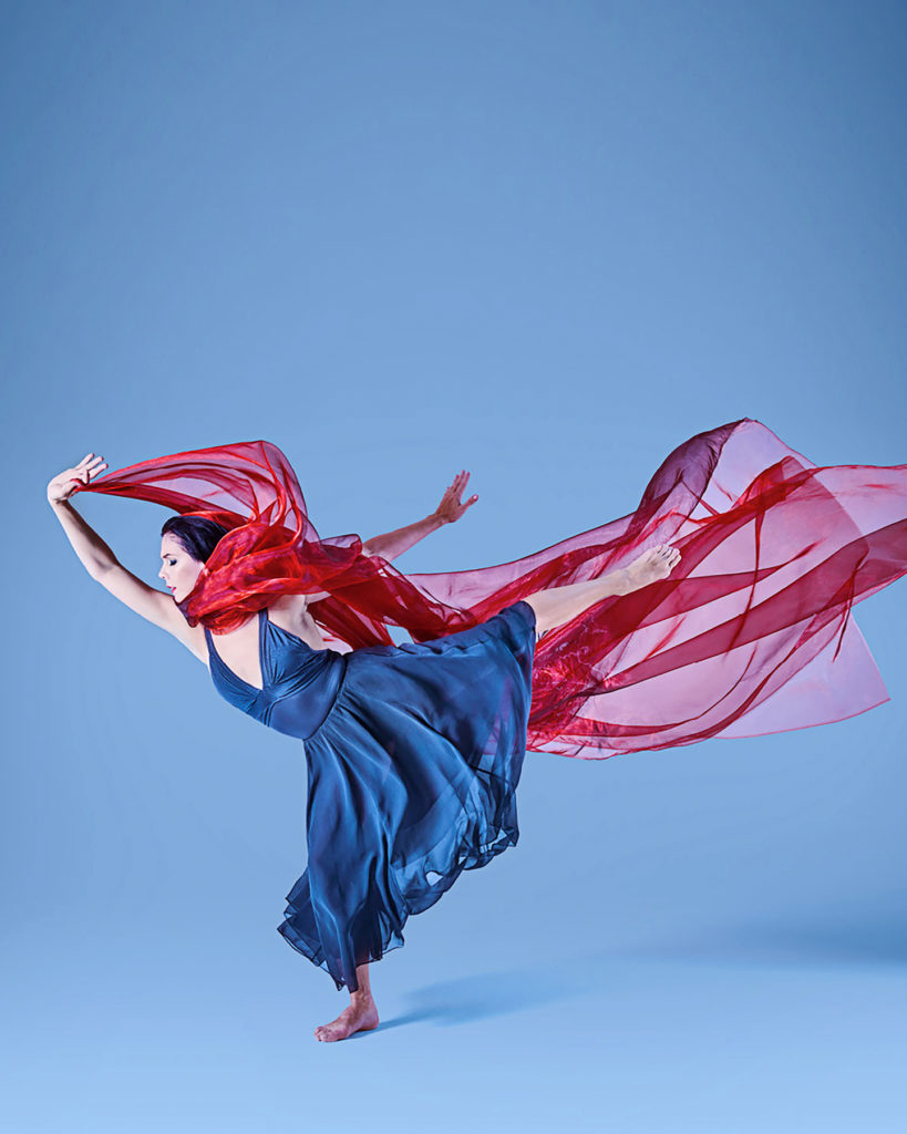 Natalia Osipova wears a flowing blue dress, holding a long stretch of red fabric that wraps around her neck and flows behind her. She poses in plié, her back leg extended long behind her, barefoot.