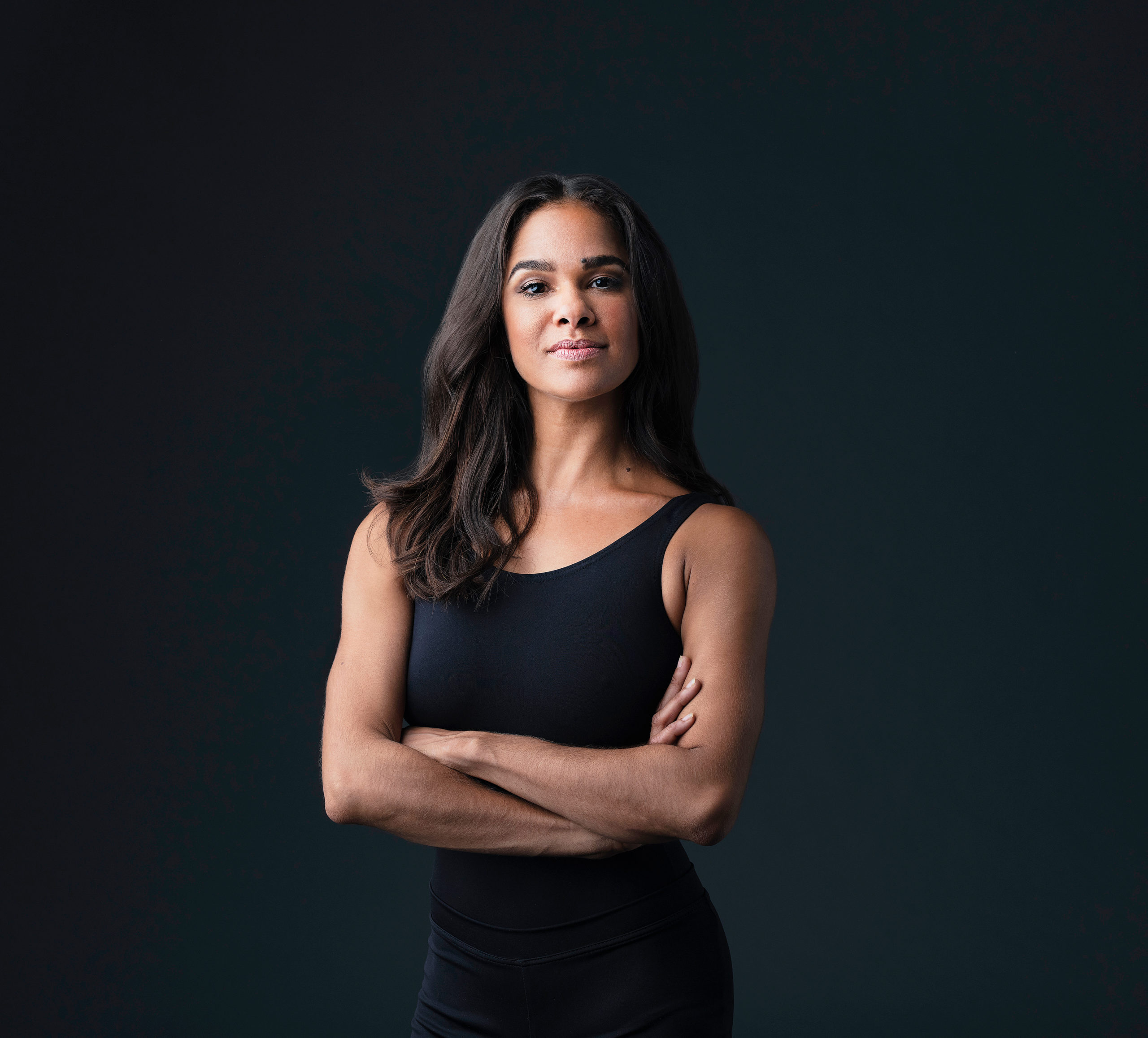 Misty Copeland poses against a black backdrop. Her arms are crossed as she looks straight at the camera with a small close-lipped smile.