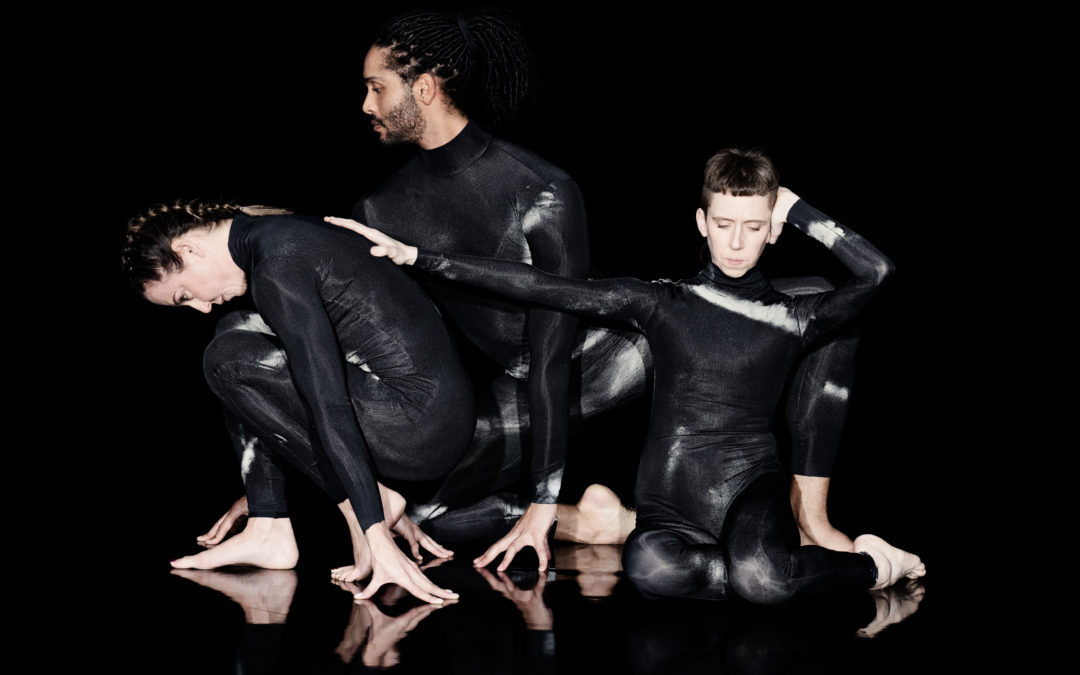 Three figures are crouched against a black background in a row wearing full length black leotards with white smoke-effect detailing. On the right is Jules Cunningham, facing forward and knelt down. They have their hair clipped short on the sides and long on the top and back. Their left hand is held behind their back and the right reaches out and rests on Melanie C’s back. In the centre is Harry Alexander, also kneeling and facing front on with his eyes gazing to the side. His hair is sectioned into locs and pulled back, and he has dark stubble on his jaw and chin. On the left is Melanie C, crouched profile and facing away from Jules. She is resting on her palms and feet with her back arched and head hanging toward the ground. Her hair is neatly plaited into a single Dutch braid.