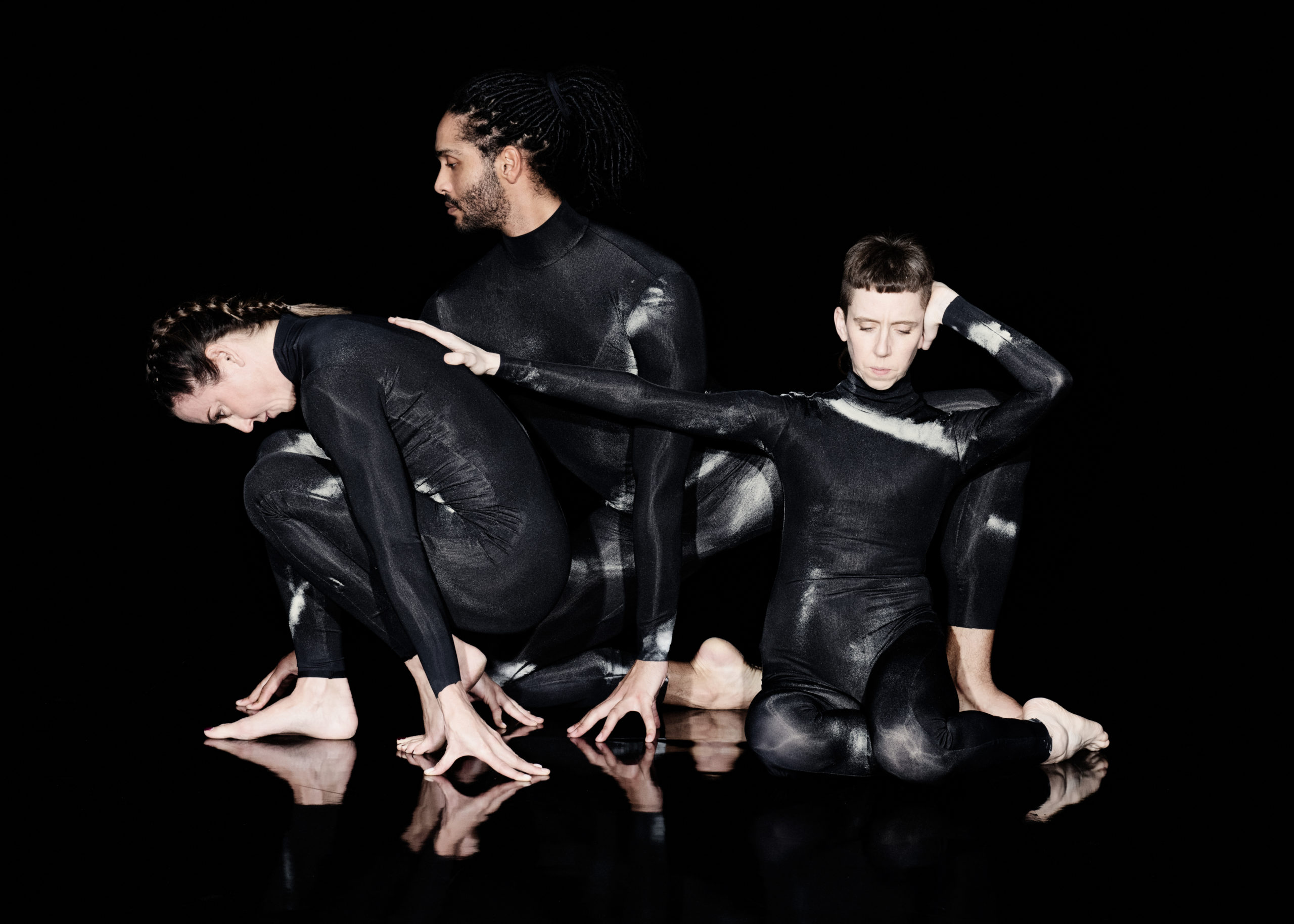 Three figures are crouched against a black background in a row wearing full length black leotards with white smoke-effect detailing. On the right is Jules Cunningham, facing forward and knelt down. They have their hair clipped short on the sides and long on the top and back. Their left hand is held behind their back and the right reaches out and rests on Melanie C’s back. In the centre is Harry Alexander, also kneeling and facing front on with his eyes gazing to the side. His hair is sectioned into locs and pulled back, and he has dark stubble on his jaw and chin. On the left is Melanie C, crouched profile and facing away from Jules. She is resting on her palms and feet with her back arched and head hanging toward the ground. Her hair is neatly plaited into a single Dutch braid.