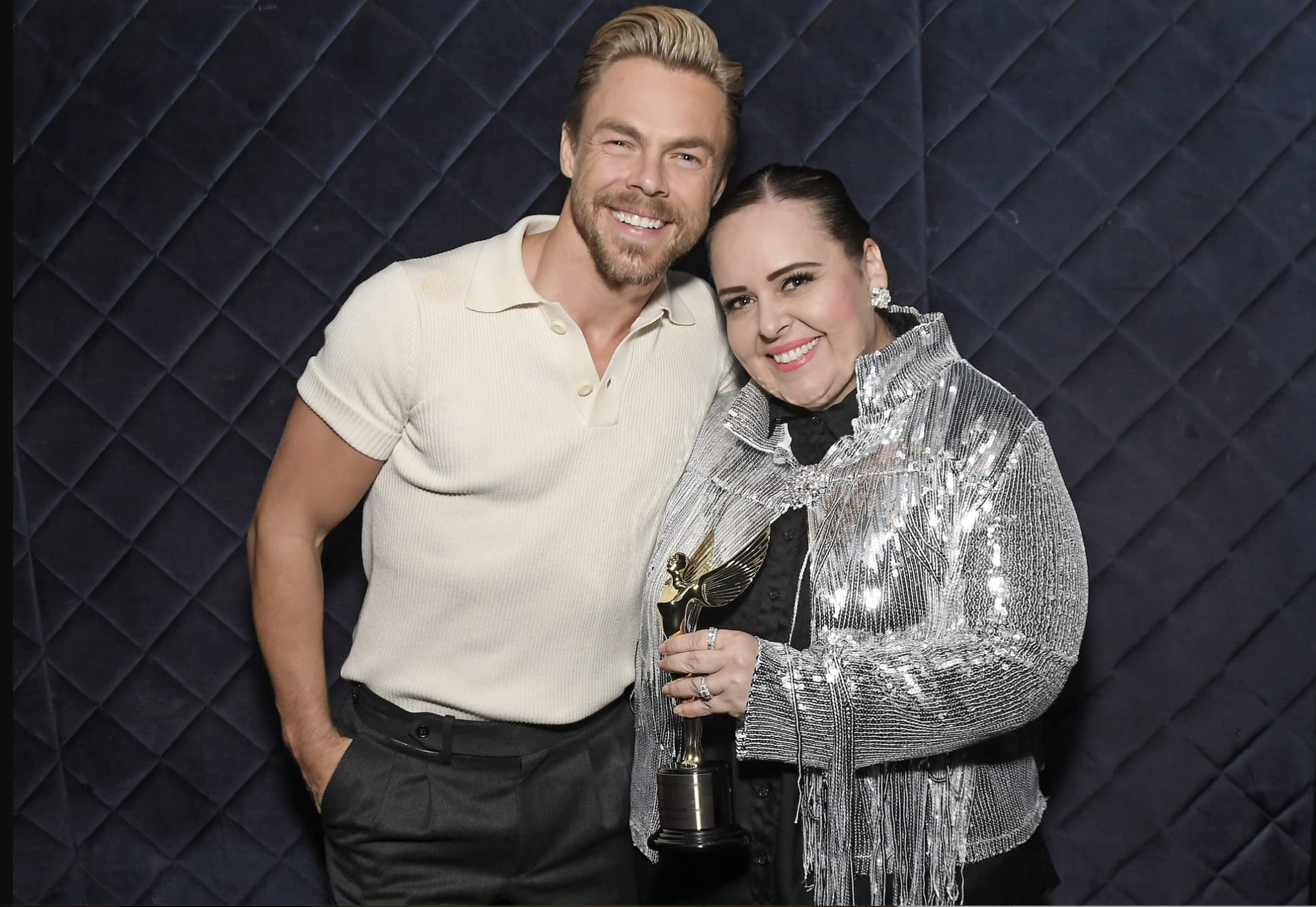 A man in a white shirt stands with a woman in a sparkly silver coat who is holding an award statuette.