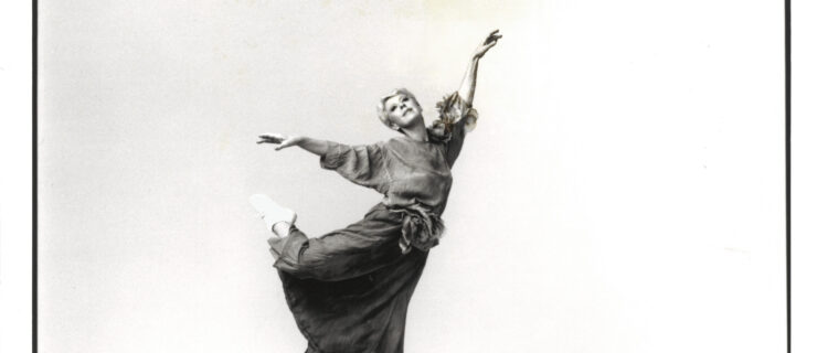 In a black and white archival image, Valda Setterfield serenely balances on one leg while the other kicks up behind her in a casual attitude. Her arms are extended, upper body light as she tips forward off of relevé. She wears chunky tennis shoes and an ankle length dress with draping sleeves.