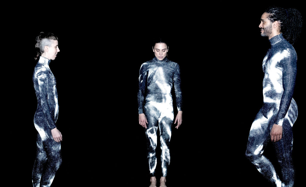 Three dancers in matching unitards that appear as textured, molten silver in the light pose against a black backdrop. One faces forward, eyes downturned, while the other two are to either side, facing the center and smiling slightly.