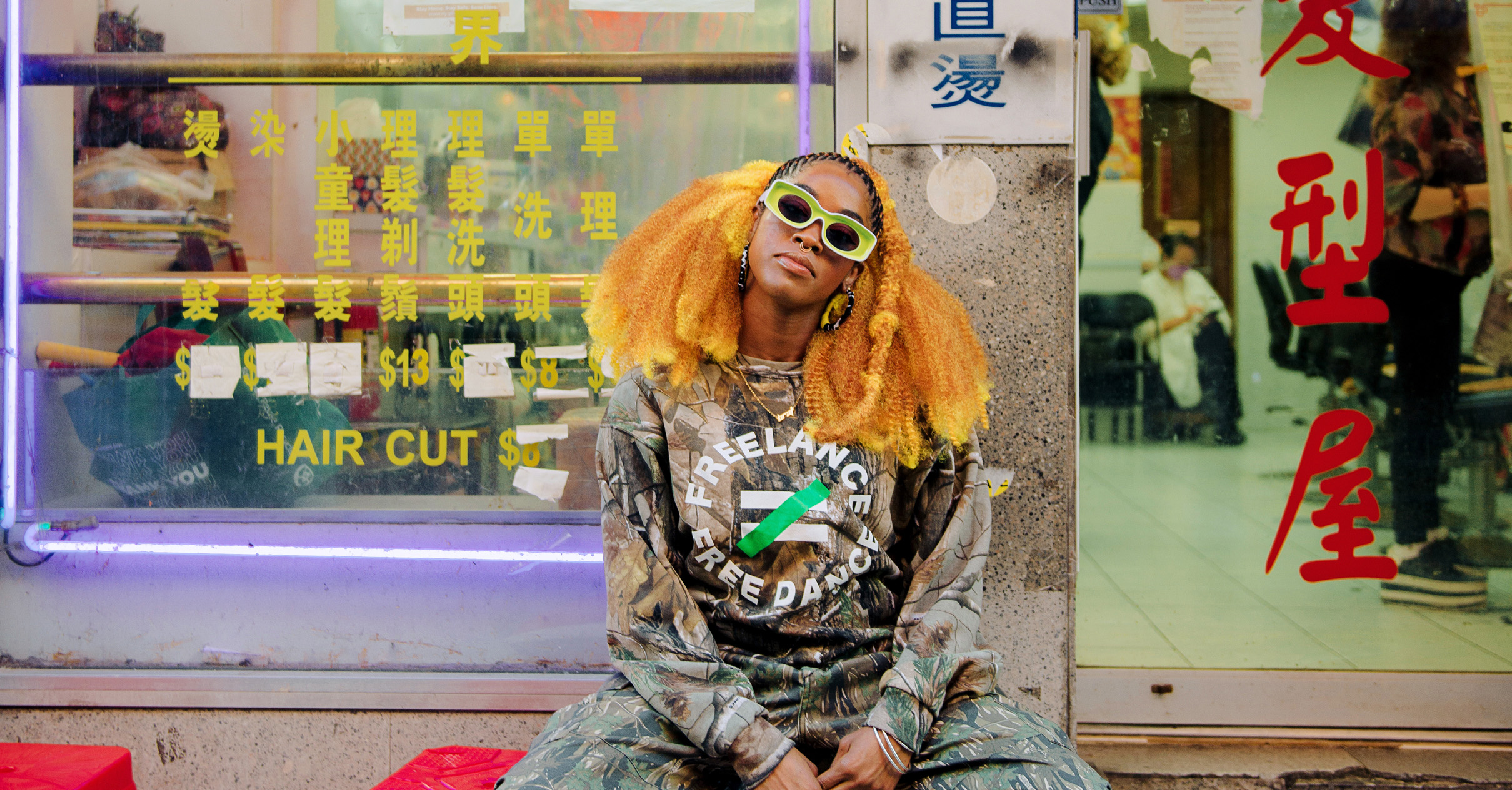 BRAT sits outside a shop in Chinatown, hands clasped between her splayed knees and head tilted to one side. Her eyes are hidden behind green sunglasses; her big, fluffy hair is a vibrant orange and reaches her shoulders.
