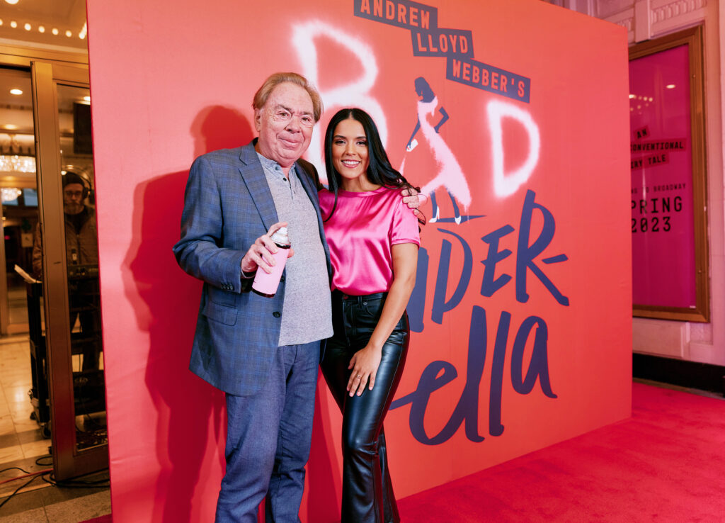 Sir Andrew Lloyd Webber holds a pink can of hairspray with one hand, while his free arm wraps around Linedy Genao, who smiles at the camera. They are posed against a red poster with a title treatment reading "Andrew Lloyd Webber's Bad Cinderella."