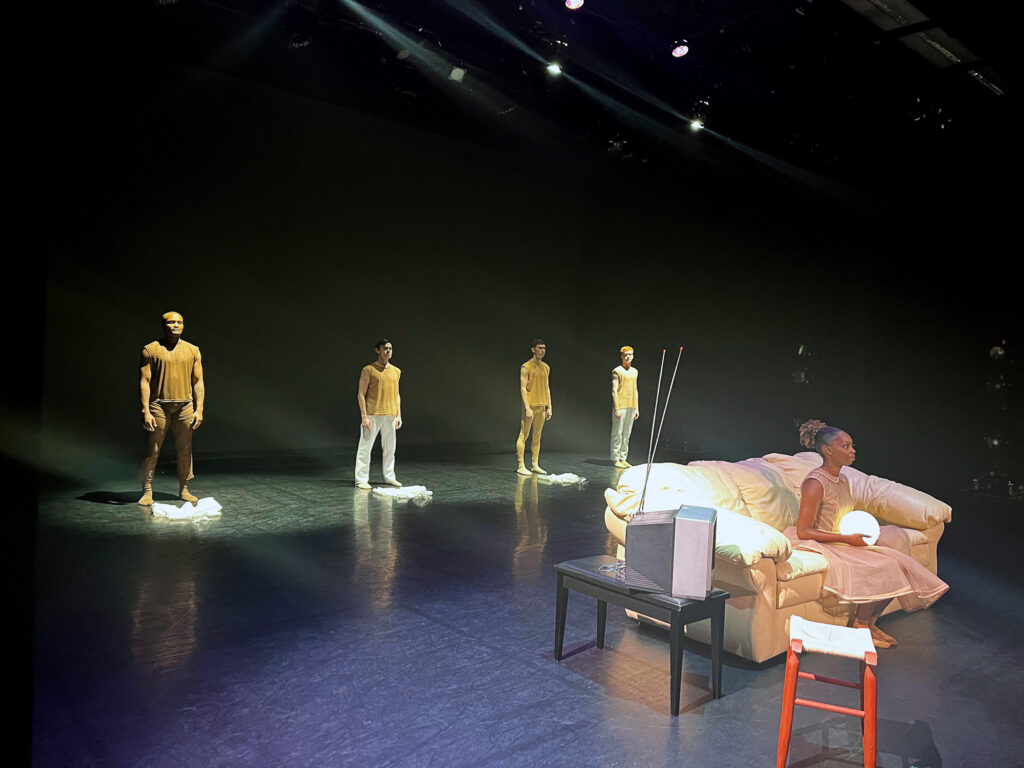 Downstage, a Black woman sits on a couch holding a glowing orb in her lap. To her right is a side table with an old-looking television. Upstage, four male dancers in yellow shirts stand in a line, facing the audience.