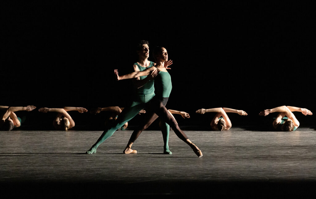 Chyrstyn Fentroy sways forward off her center of balance as her front foot skims off the floor. Her arms are extended straight to the side, hands flexed, as her partner lunges behind her to support her with hands under her shoulders. They are costumed in green and black; Chyrstyn's shoes match her skin tone. Upstage, a line of dancers lie on their backs, arms crossed at the elbows.