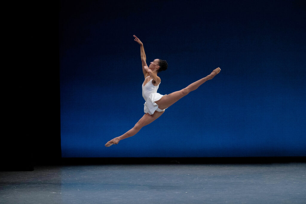Chyrstyn Fentroy is caught midair, dancing alone onstage. Her legs are in a 180 degree split, back leg higher than the front, her front arm extended high in front of her. She smiles, chin raised to follow her top hand. She wears a white leotard with an attached short skirt over tights and pointe shoes that match her skin tone.