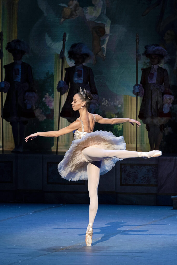 Onstage, Chyrstyn Fentroy balances in attitude back en pointe, angled to face an upstage diagonal. She smiles serenely as she gazes past her front arm, gently raised in second arabesque. She is costumed in pink tights and pointe shoes and a champagne colored classical tutu.