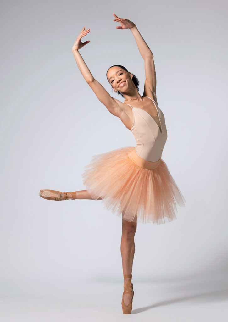 Chyrstyn Fentroy smiles widely at the camera as she poses in croisé attitude back, arms in high fifth as she sways toward her supporting leg. Her shoes are pancaked to match her skin. She wears a fluffy, muted orange practice tutu over a pale leotard.