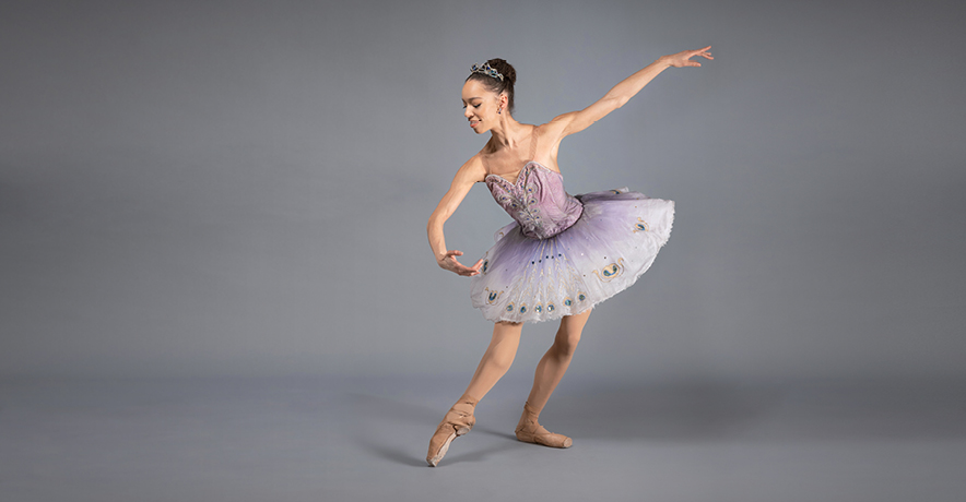 Chyrstyn Fentroy poses in tendu effacé devant, standing leg in plié. Her gaze is downturned as her épaulement shifts her torso over her working leg, downstage arm extending elegant and long beside her. She wears a pale purple classical tutu over tights and pointe shoes that match her skin tone.