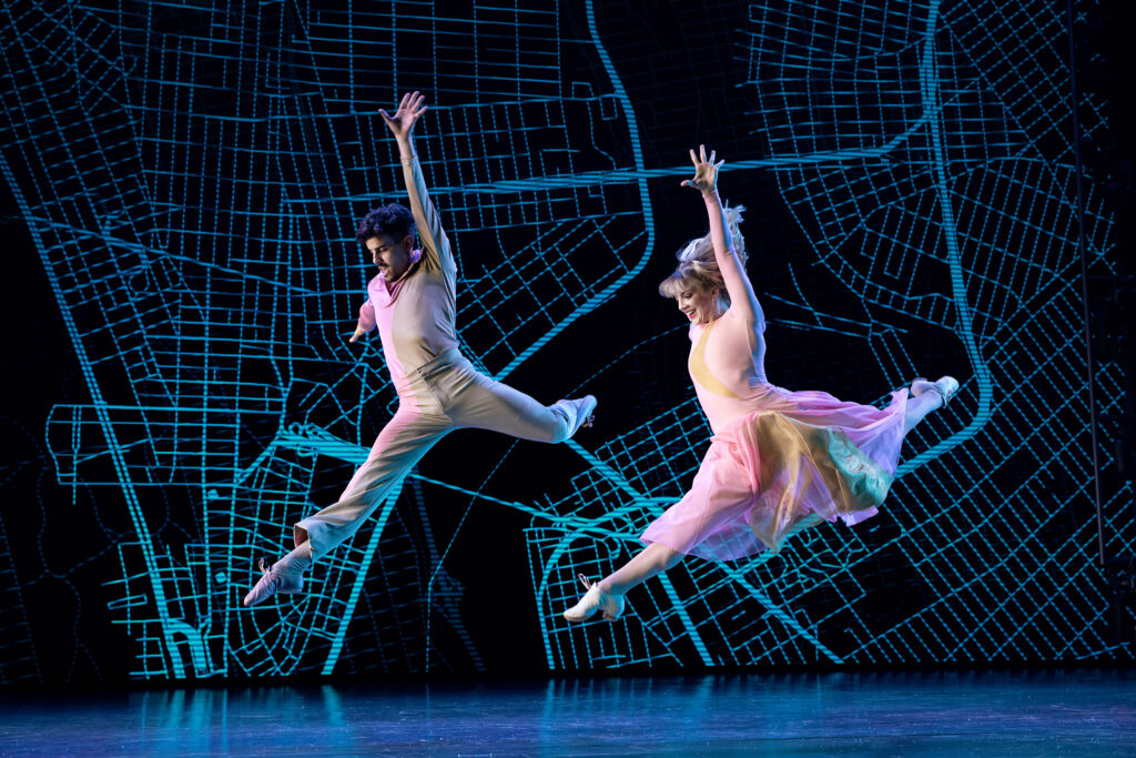 Two dancers are caught mid-leap onstage, back legs bent in attitude. Their downstage arms reach with open palms overhead, while they gaze past their front legs with exhilarated smiles. The woman wears a flowing pink dress, the main khakis and a long sleeve shirt. The backdrop shops a blue grid pattern recognizable as a map of New York City.
