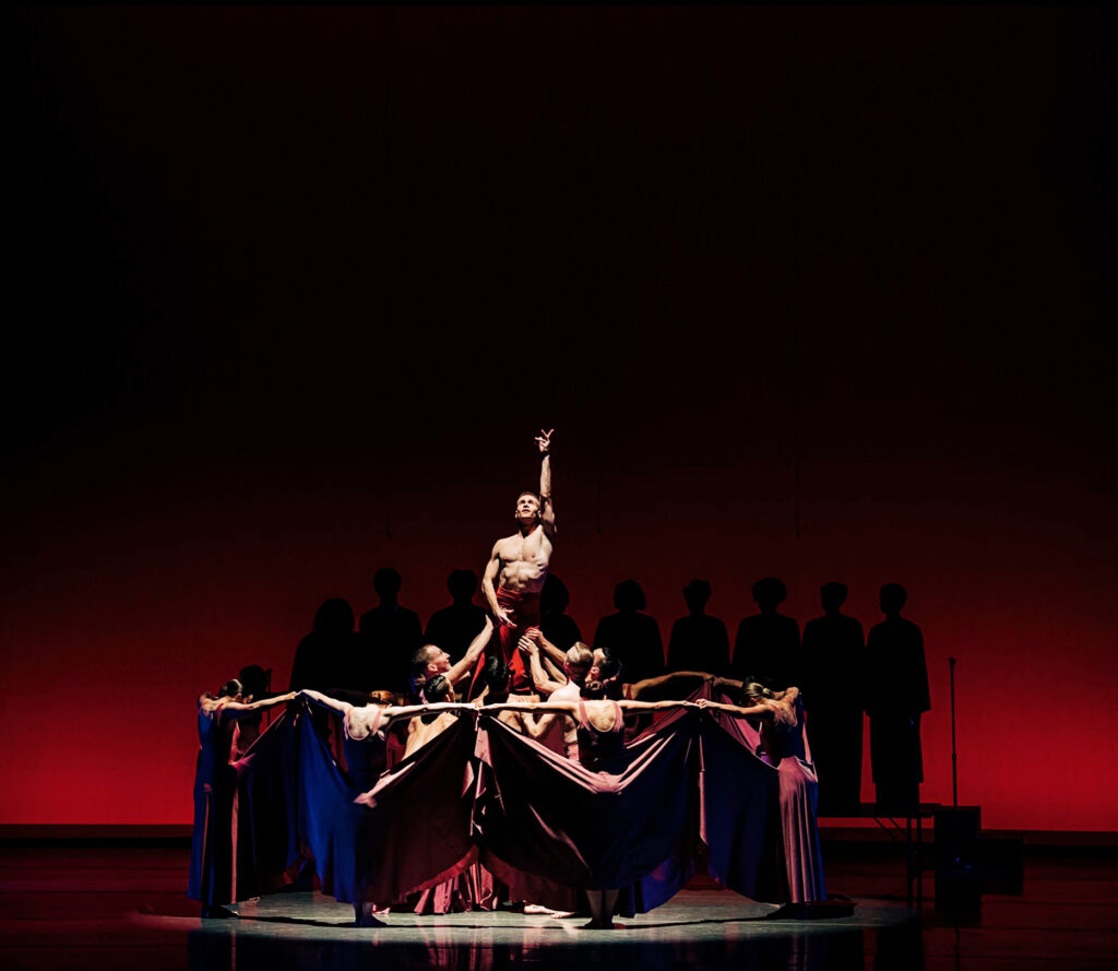 On a shadowy stage, a shirtless male dancer is lifted from the center of a cluster as he reaches one arm to the sky. A half dozen dancers form a circle around the cluster, pulling their long skirts up and to the sides to create a barrier. A line of silhouetted figures are visible upstage on a riser.