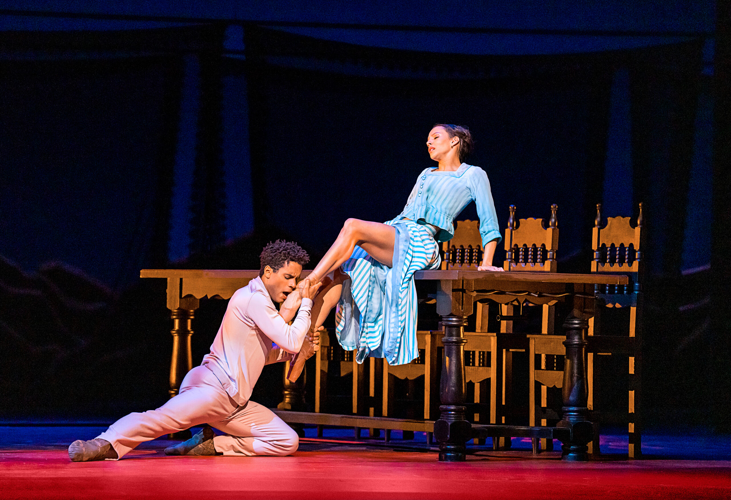 Marcelino Sambé and Francesca Hayward in performance. Sambé kneels as he desperately cradles one of Hayward's feet to his face. Hayward, seated on a kitchen table, leans back on her hands, eyes closed in tension and pleasure. Her long, blue and white striped skirt slips up her thighs.