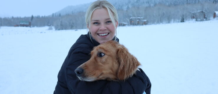 Haley Hilton, blond in a black jacket, hugs her brown dog in front of a snowy wilderness background.