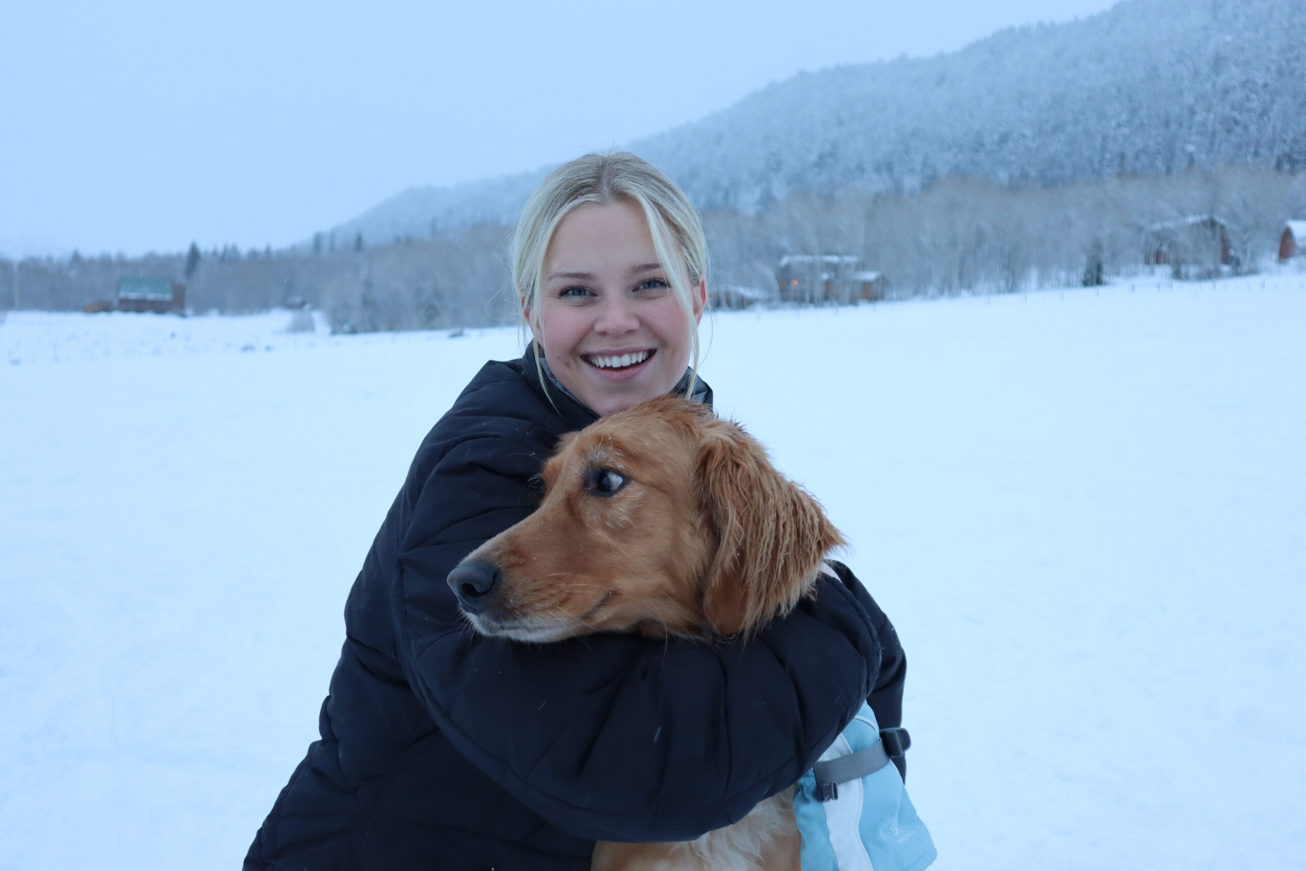 Haley Hilton, blond in a black jacket, hugs her brown dog in front of a snowy wilderness background.