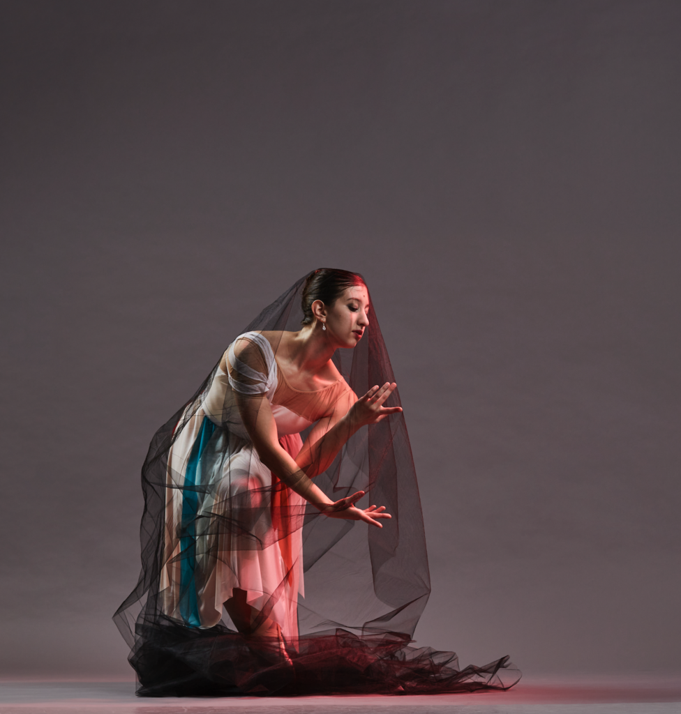 A dancer in a black mesh veil draped over her head and the long white dress she wears poses on a grey backdrop. She pliés and leans forward to twist over one leg, hands upturned and curving toward her torso as though gathering something to her.