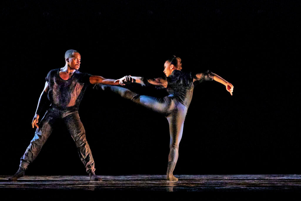 a female and male dancer wearing all black and holding hands, female has one leg extended 