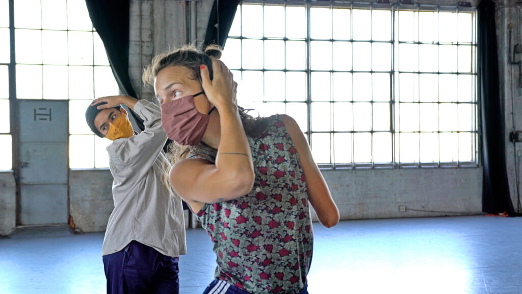 two dancers in a studio wearing masks and holding their heads with one hand