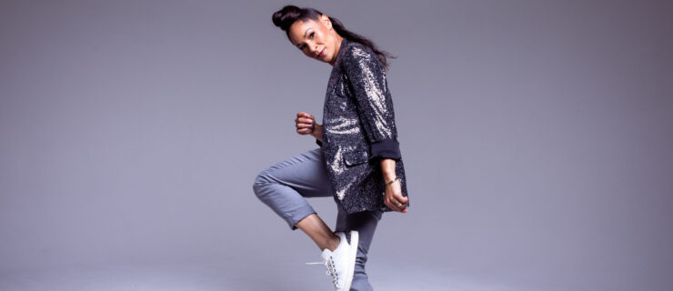 Ayodele Casel poses against a grey backdrop. In sneakers, she balances in a forced arch parallel retiré facing side. She leans her head forward to gaze seriously at the camera, her upstage arm rising to her ribs. She wears cuffed jeans and a sparkly grey blazer; her hair is piled high atop her head and tumbles in a long ponytail down her neck.