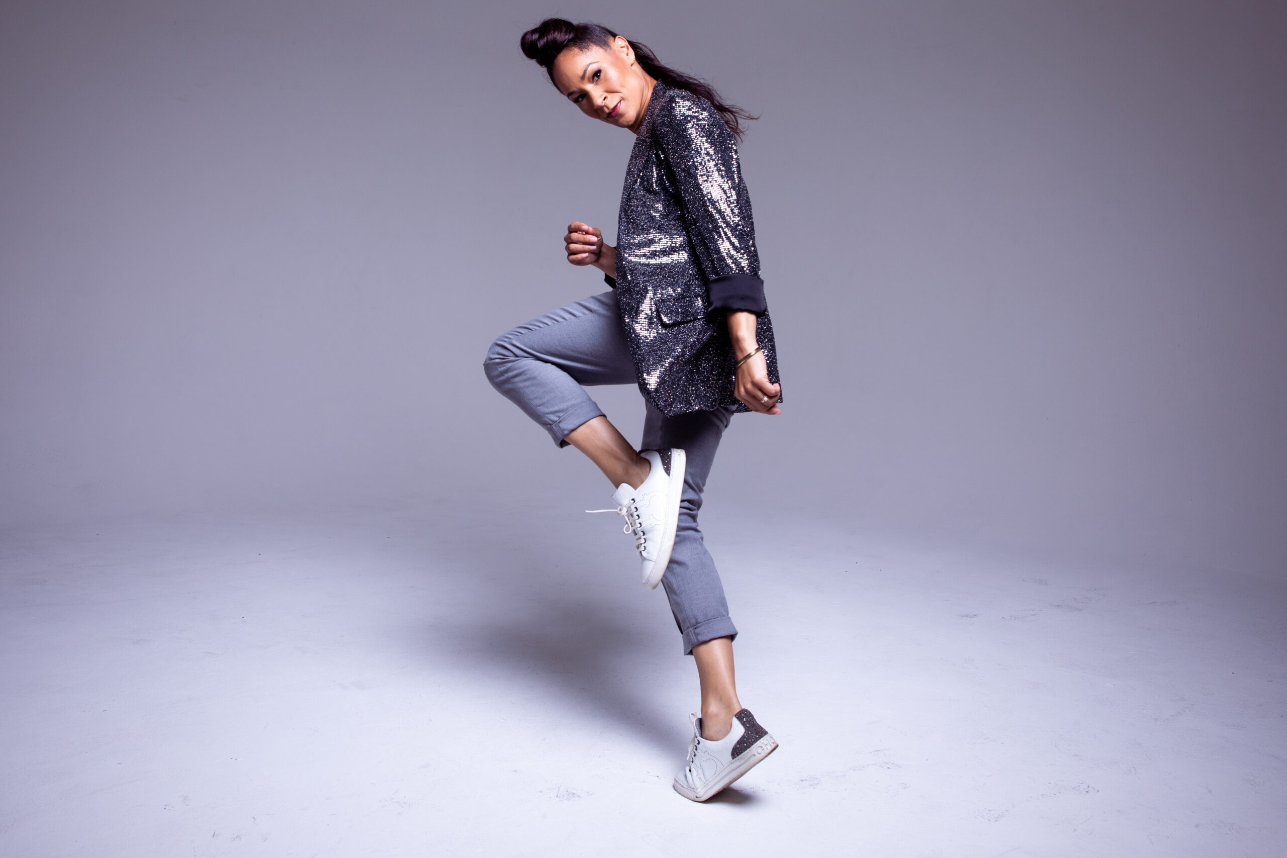 Ayodele Casel poses against a grey backdrop. In sneakers, she balances in a forced arch parallel retiré facing side. She leans her head forward to gaze seriously at the camera, her upstage arm rising to her ribs. She wears cuffed jeans and a sparkly grey blazer; her hair is piled high atop her head and tumbles in a long ponytail down her neck.