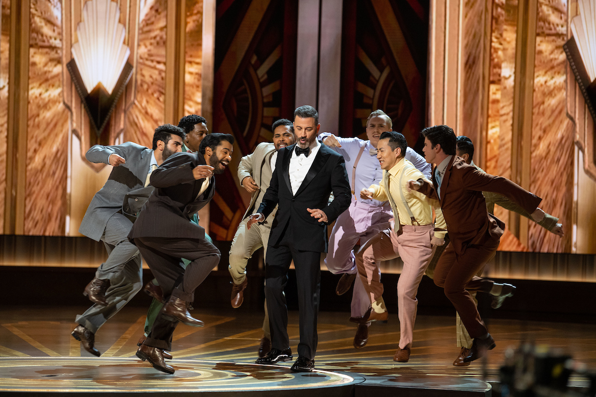 On the gilded Oscars stage, a group of eight male dancers in suits surround Jimmy Kimmel, who wears a black tuxedo, as they energetically perform the “Naatu Naatu” hook-step dance move. Kimmel smiles and looks down as he dances along.