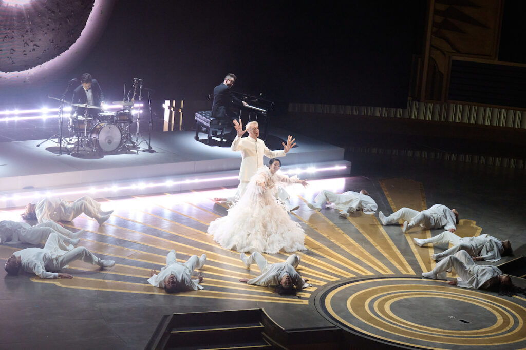 

Onstage at the Oscars awards ceremony, Stephanie Hsu wears an extravagant feathered white dress as she sings alongside David Byrne, who wears a white modern-styled suit and “hot dog” fingers. Around them on the floor in a circle, an ensemble of ten dancers in white costumes reminiscent of martial arts uniforms do a contraction on their backs. A group of three male musicians plays at the back of the stage, behind them a projection of a large black space-like ring.
