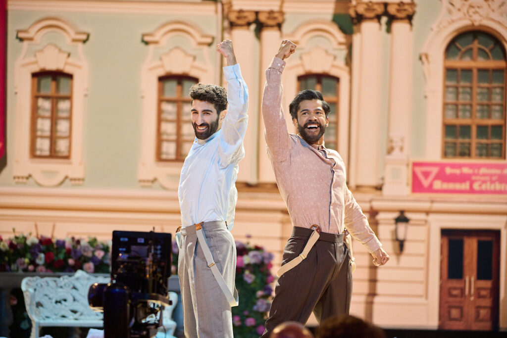 On the Oscars stage, two male South Asian dancers pose triumphantly, fists raised together, and smile jubilantly in the final pose of an energetic dance number. They wear dress shirts with suspenders and pants and stand in front of a backdrop colonial-esque architecture.