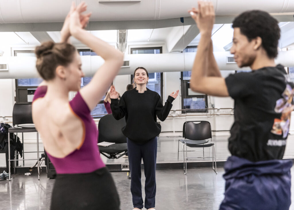 male and female dancers stand near the camera with arms up, female choreographer stands near mirror smiling 