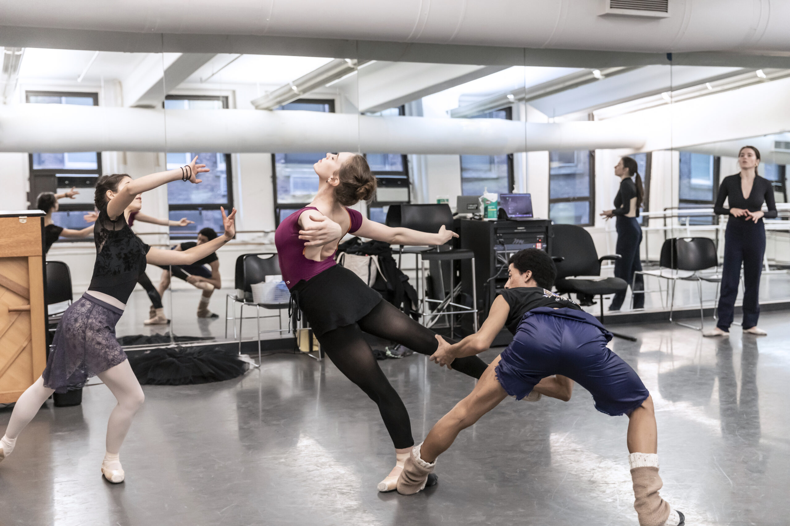 three dancers in the studio: one female learning back in arabesque, a male dancer supporting her leg, and another female reaching towards them in a lunge