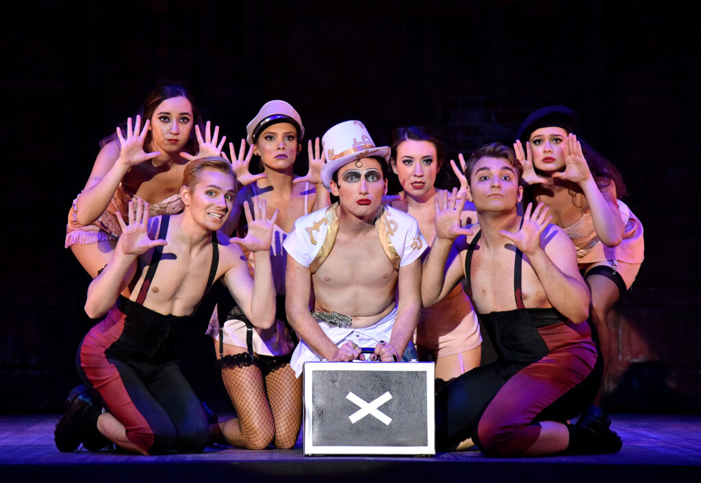 seven performers kneeling downstage, one wearing a white top hat, the others holding up their hands