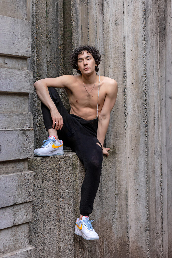 shirtless male dancer sitting on cement wall wearing black pants and nike shoes