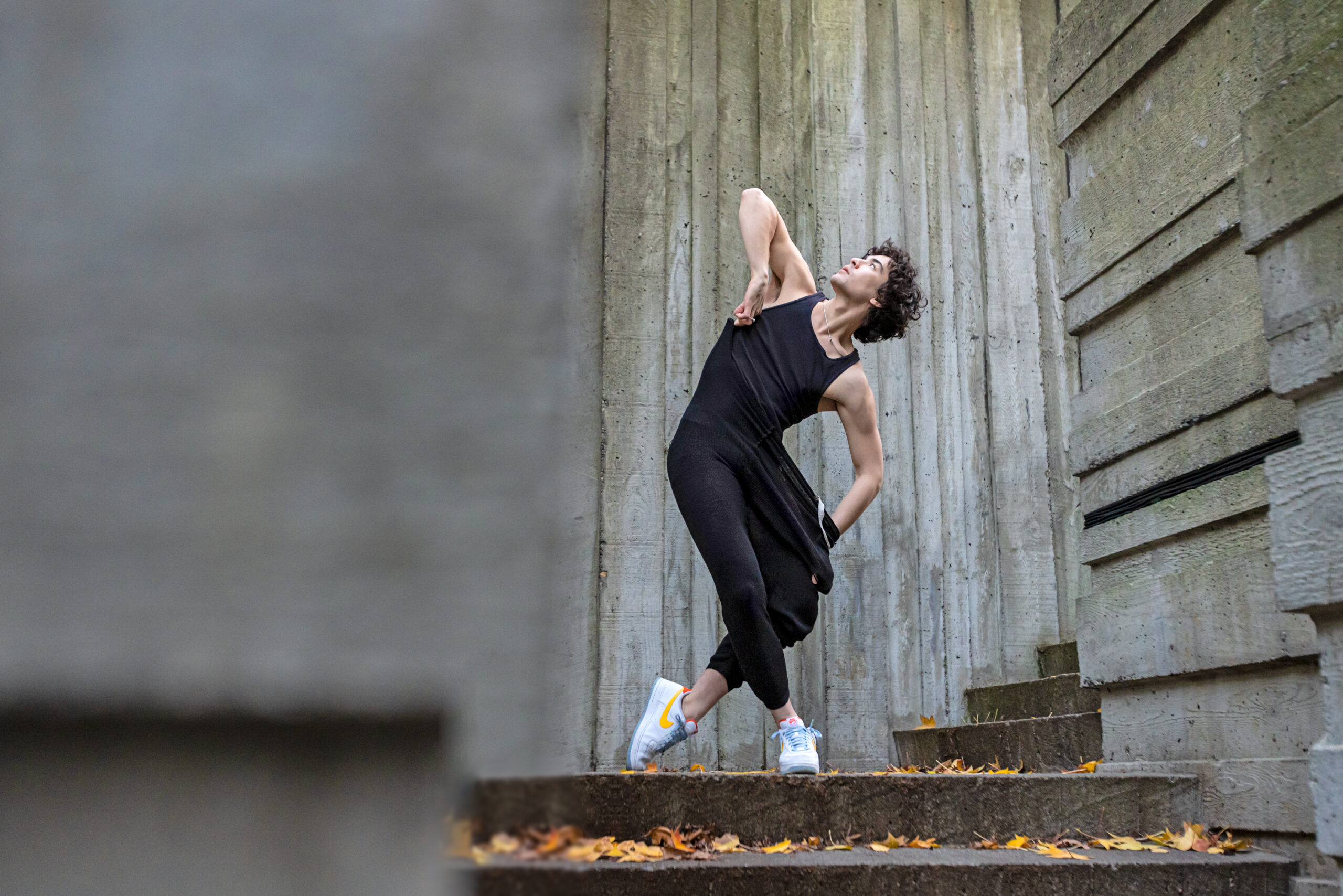 male dancer wearing all black and nike sneaker leaning laterally next to a cement wall