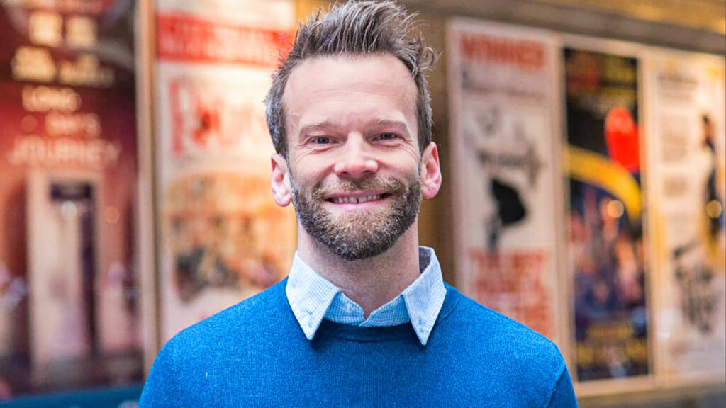 male wearing a button down and blue sweater smiling at the camera