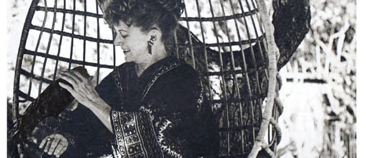 In a black and white archival image, Mia Slavenska, here in her 50s, sits in an egg-shaped, woven chair outside. Perched on her wrist is a black crow, who she gently pets with her free hand as she smiles down at it. She wears a thick robe or dress with geometric patterns; her curly hair is piled atop her head.