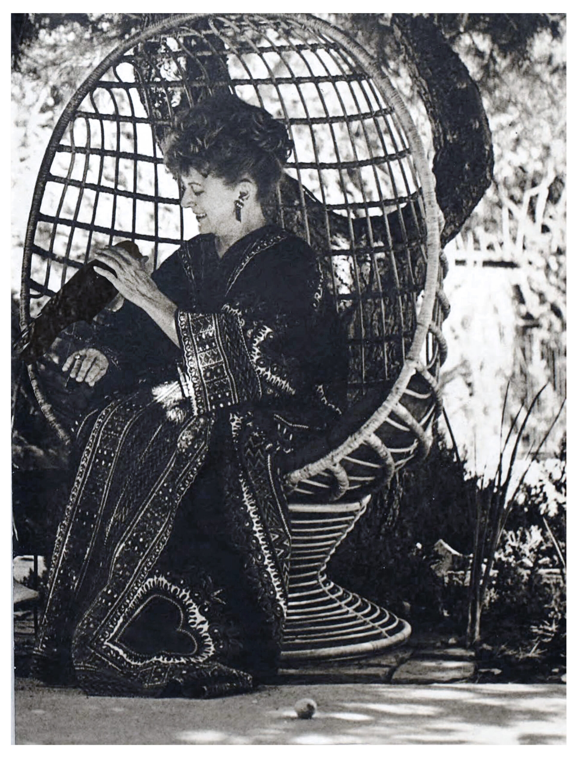 In a black and white archival image, Mia Slavenska, here in her 50s, sits in an egg-shaped, woven chair outside. Perched on her wrist is a black crow, who she gently pets with her free hand as she smiles down at it. She wears a thick robe or dress with geometric patterns; her curly hair is piled atop her head.
