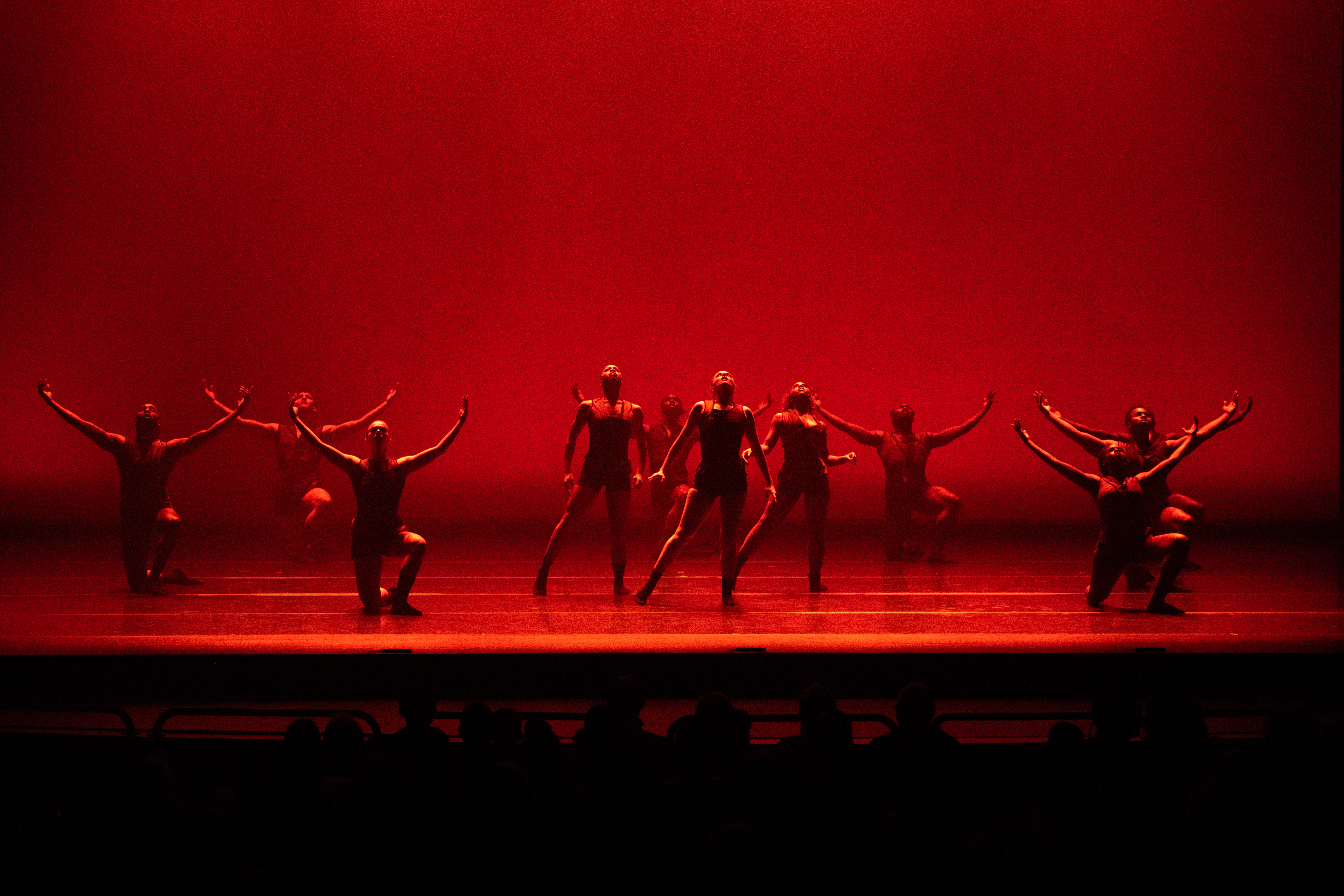 Clusters of dancers appears on a stage awash in red, their faces falling into shadow. A trio at the center stands leaning into one hip, while encircling them are other dancers on one knee, facing front, their arms curving up from their shoulders.