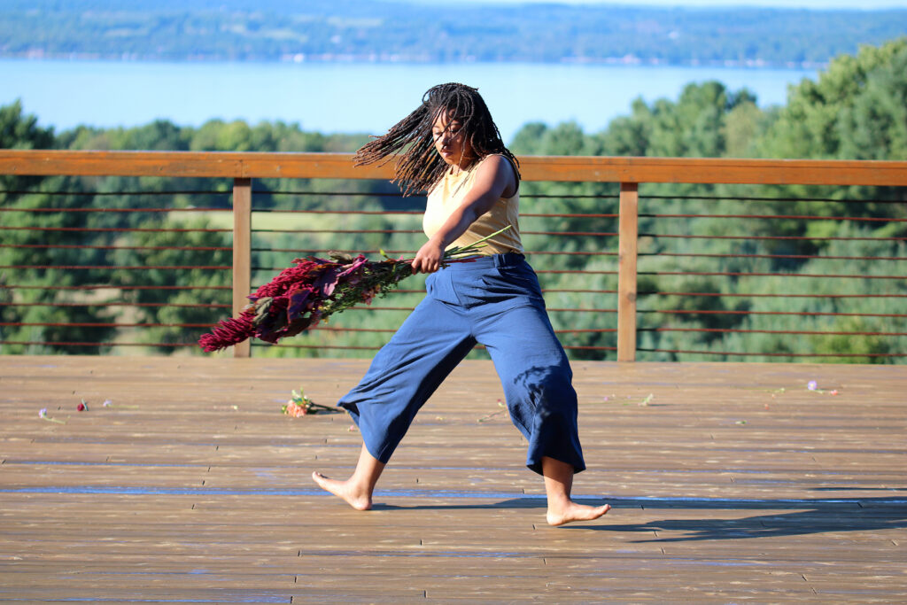 female dancer wearing flowy blue pants dancing with a bundle of flowers on a deck